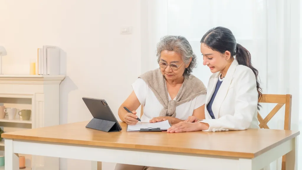 Two women, one elderly and one younger, seated at a wooden table with a tablet, reviewing documents together.