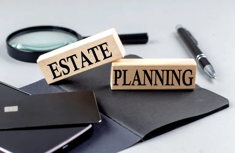 Wooden blocks with "ESTATE" and "PLANNING" text, black pen, magnifying glass, and credit card on notebook.