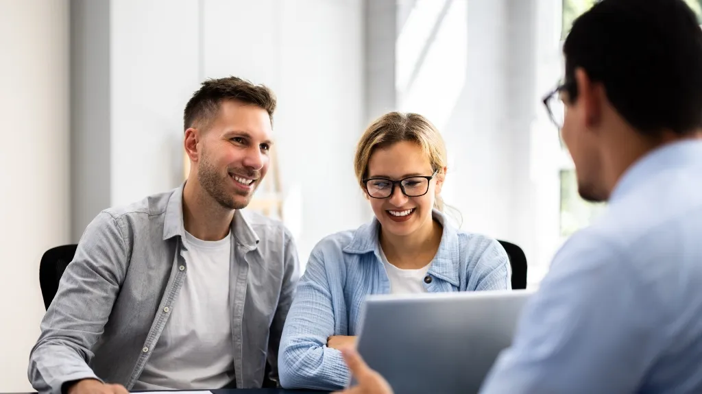 Three professionals, two women and one man, engaged in a discussion around a laptop in a bright office.
