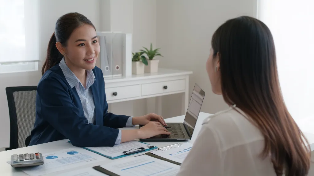 Two professional women engaged in a discussion at a desk with documents and a laptop.