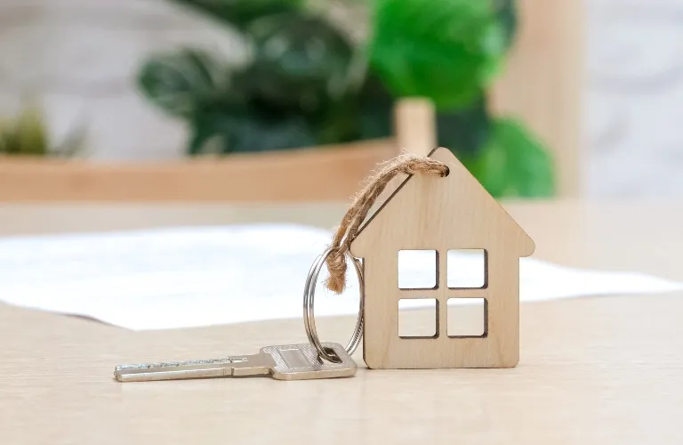 Silver house key attached to a wooden house-shaped keychain on a light wooden surface.