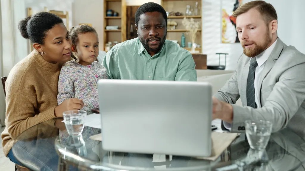 A professional man in a suit explaining information on a laptop to a family of three at a glass table.