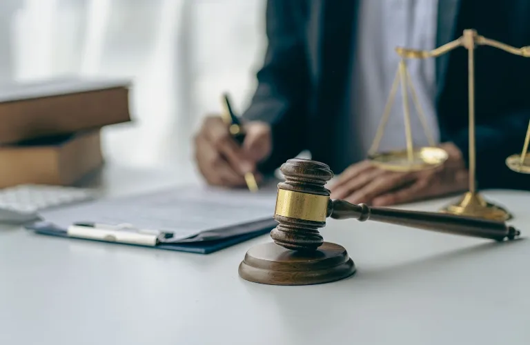 Wooden judge's gavel and brass scales of justice on white desk with person writing.