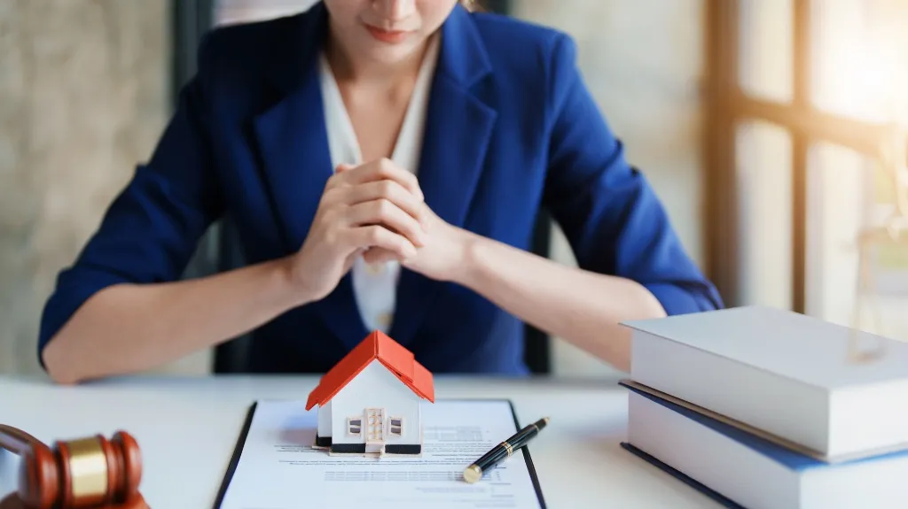 Professional woman in blue blazer with miniature house model, documents, and gavel on desk.