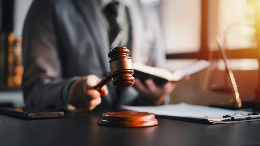 Close-up of a judge’s hand holding a wooden gavel above a sound block in a courtroom setting.